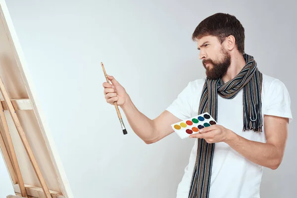 A man artist paints on an easel a brush and paint in the hands of a hobby creativity light background — Stock Photo, Image