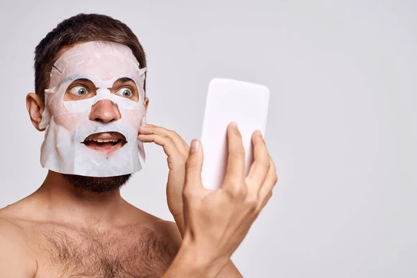 Un homme avec un masque nettoyant sur le visage se regarde dans un miroir sur un fond clair — Photo