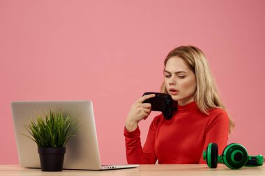 Dizüstü bilgisayarın önündeki masada oturan bir kadın elinde bir oyun platformuyla teknoloji kulaklığı çalıyor. 