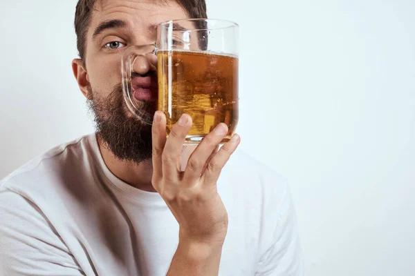 Hafif arka planda bira fincanı olan sakallı bir adam. Beyaz tişörtlü, alkollü bir içkinin görüntüsü. — Stok fotoğraf