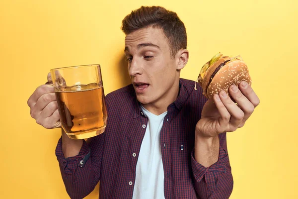 Homme joyeux avec une tasse de bière et un hamburger sur ses mains régime alimentaire style de vie jaune fond — Photo