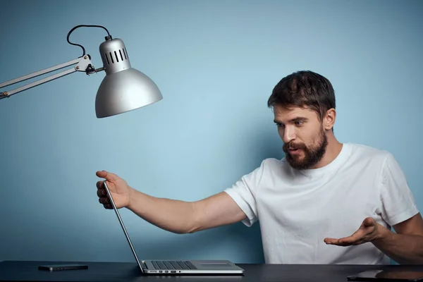 Um homem com um laptop dentro de casa em uma mesa em um fundo azul e escritório de trabalho lâmpada — Fotografia de Stock