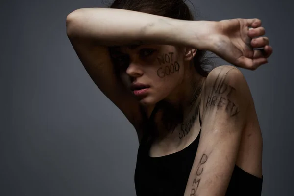 Mulher frustrada com inscrições ofensivas em seu corpo e em uma camiseta preta em um retrato de fundo cinza — Fotografia de Stock
