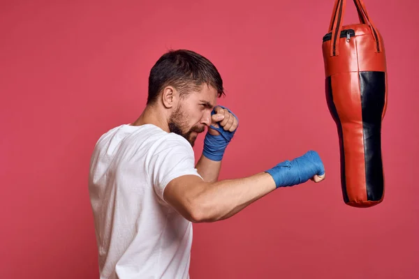 Un uomo vicino a un sacco da boxe con guanti blu e una maglietta bianca sta praticando pugni sportivi — Foto Stock