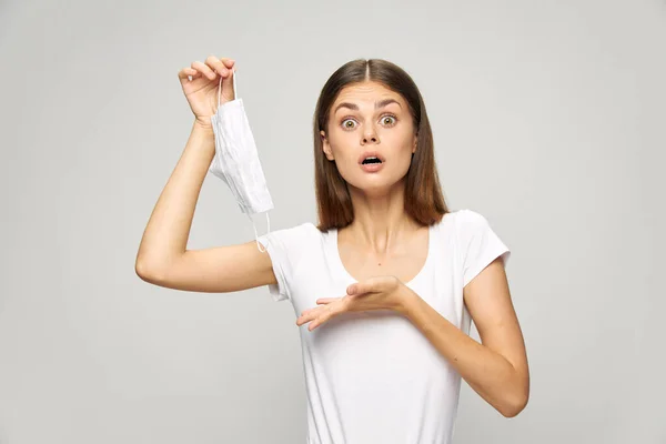 Mulher com uma expressão surpresa em seu rosto com uma máscara médica em sua mão t-shirt branca — Fotografia de Stock