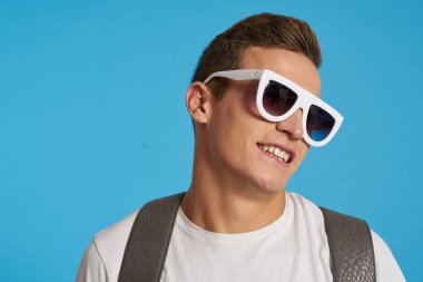 Kotlu, dizleri yırtık rahat bir adam mavi arka planda bir sandalyede oturuyor ve yüzünde uzun gözlüklerle oturuyor.