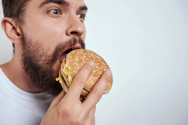 白いTシャツに身を包んだ男がハンバーガーを食べると、空腹のファーストフードが近くに見える — ストック写真