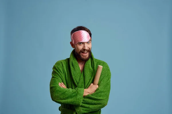 A man with a pink sleep mask holds a rolling pin in his hand and a green robe blue background emotions model — Stock Photo, Image