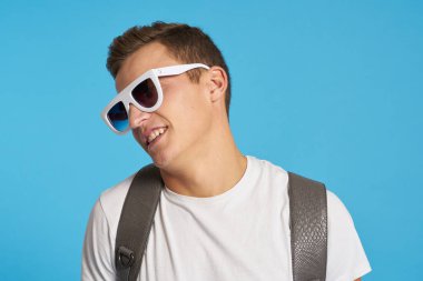 Kotlu, dizleri yırtık rahat bir adam mavi arka planda bir sandalyede oturuyor ve yüzünde uzun gözlüklerle oturuyor.