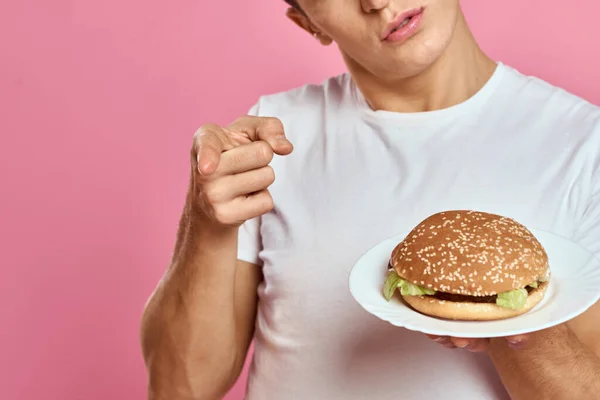 Emocionální chlap s hamburgerem na talíři a bílé tričko růžové pozadí oříznutý pohled na kalorie rychlého občerstvení — Stock fotografie