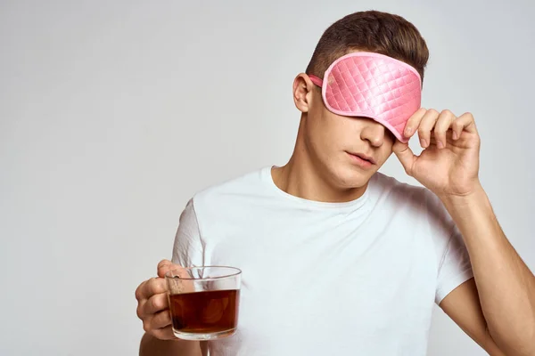 Homem bonito com máscara de sono rosa e com uma xícara de chá na mão puxa-se sobre um fundo leve — Fotografia de Stock