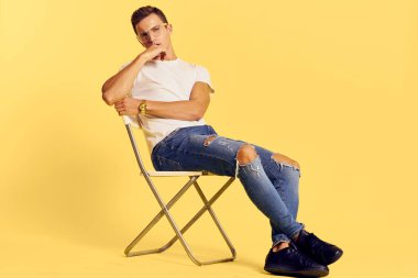 Sandalyede oturan yakışıklı adam beyaz tişört kot pantolon modern tarz sarı arka plan