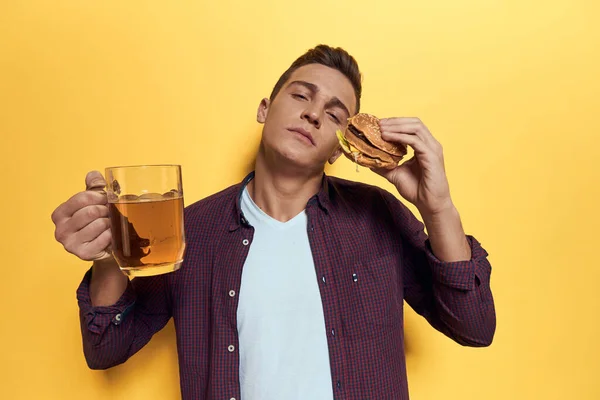 Homme joyeux avec une tasse de bière et un hamburger sur ses mains régime alimentaire style de vie jaune fond — Photo