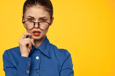 Mavi gömlekli, gözlüklü, yüzünde parlak makyajlı bir kadın elleriyle işaret ediyor.