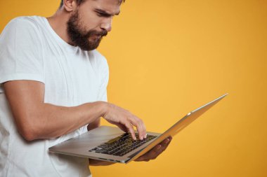 Sakallı bir adam elinde dizüstü bilgisayarla sarı arka plan klavyesinde yeni teknolojileri izliyor.