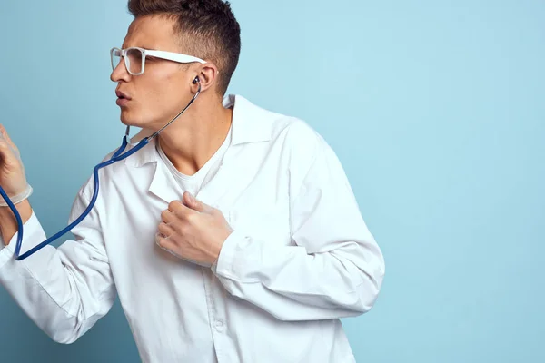 Ein Mediziner in Bademantel und Brille hält vor blauem Hintergrund ein Stethoskop in der Hand. — Stockfoto