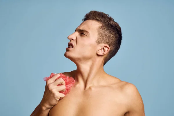 Молодой парень с розовой мочалкой в руке на синем фоне кожи уход дерматологии модели обрезанный вид — стоковое фото