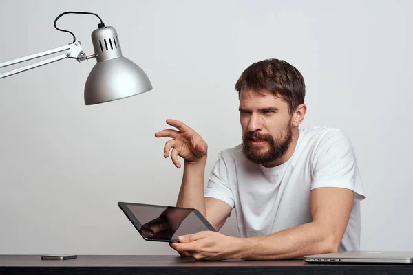 Un hombre con una tableta en la mesa hace gestos con las manos sobre un fondo claro y una lámpara de hierro — Foto de Stock