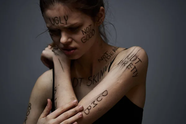 Beleidigte Frau mit wütenden Schriftzügen auf ihrem Körper berührt sich mit den Händen auf grauem Grund — Stockfoto