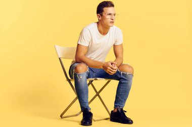 Sandalyede oturan yakışıklı adam beyaz tişört kot pantolon modern tarz sarı arka plan