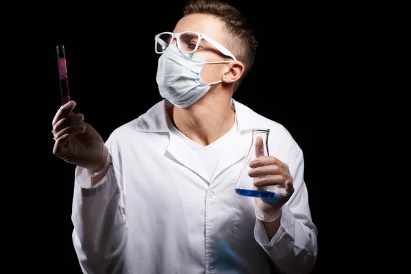 Assistant de laboratoire dans un masque médical et dans un manteau blanc avec un liquide dans une fiole et un produit chimique sur un fond noir — Photo