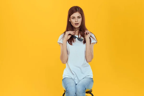 Uma mulher senta-se em uma cadeira e segura óculos escuros em suas mãos uma camiseta branca roupas femininas moda fundo amarelo — Fotografia de Stock