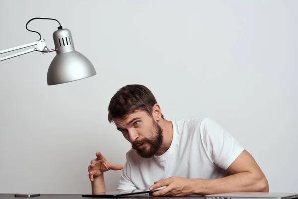 Un hombre con una tableta en la mesa hace gestos con las manos sobre un fondo claro y una lámpara de hierro — Foto de Stock