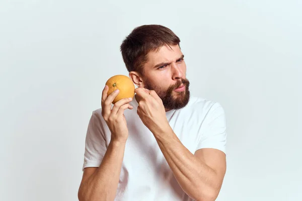 Un hombre con una naranja en una camiseta blanca sobre un fondo claro la dieta de una barba gruesa morena — Foto de Stock