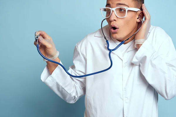 Un trabajador médico con una bata y gafas sostiene un estetoscopio en sus manos sobre un fondo azul vista recortada —  Fotos de Stock