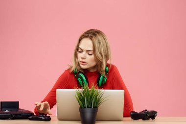 Kulaklık takan, dizüstü bilgisayarlı bir kadın masa başı eğlence sektöründe pembe arka planda oturuyor.