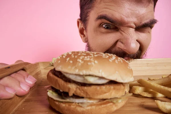 Tipo agresivo con una bandeja en sus manos hamburguesa patatas fritas fondo rosa mirada hambrienta — Foto de Stock