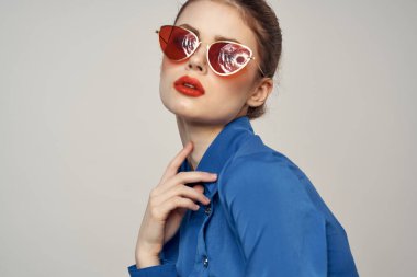 Güneş gözlüklü modaya uygun bir kadın ve elleri açık arka planda beliren mavi bir gömlek.