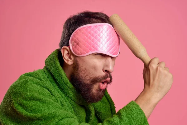 Um homem com uma máscara de dormir cor-de-rosa no rosto com um roupão verde com um rolo na mão — Fotografia de Stock
