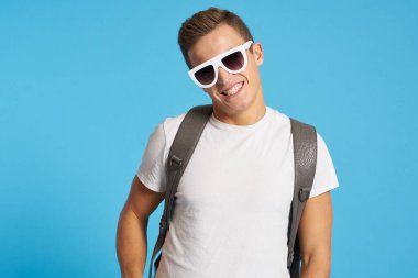 Kotlu, dizleri yırtık rahat bir adam mavi arka planda bir sandalyede oturuyor ve yüzünde uzun gözlüklerle oturuyor.