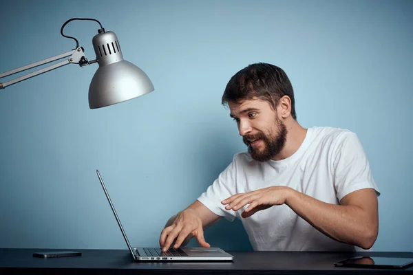 Een man met een laptop binnen aan een tafel op een blauwe achtergrond en een lampenkantoor Stockfoto