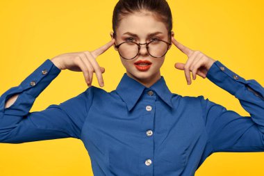 Gözlüklü ve mavi gömlekli kadın sarı arka planda el kol hareketi yapıyor.
