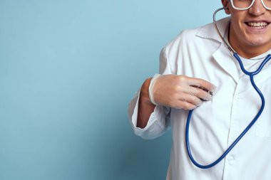 Gecelik ve gözlüklü bir sağlık çalışanı mavi arka planda steteskop tutuyor.