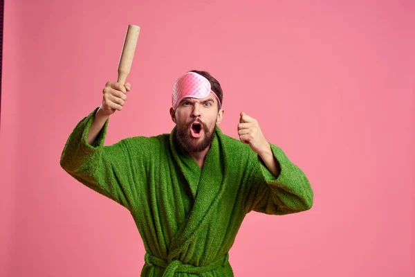Retrato de un hombre con una máscara de sueño rosa y un rodillo de madera emociones modelo de irritabilidad bata verde — Foto de Stock