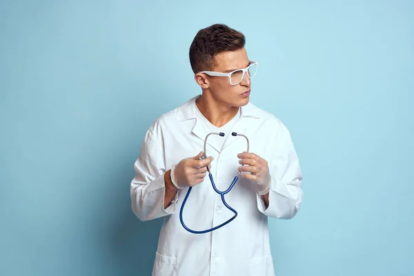 Gecelik ve gözlüklü bir sağlık çalışanı mavi arka planda steteskop tutuyor. — Stok fotoğraf