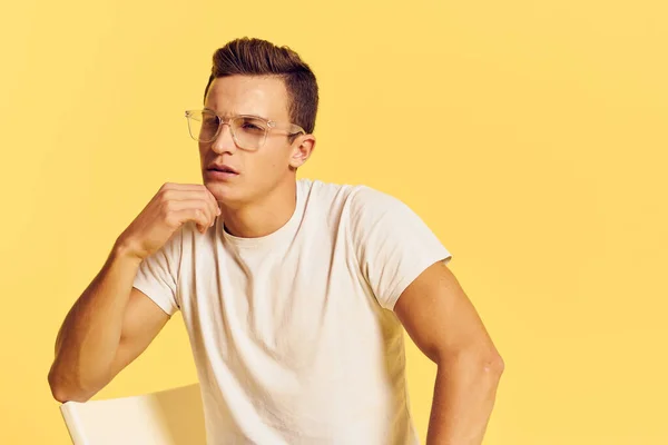 Un hombre con una camiseta blanca se sienta en una silla con gafas emoción amarillo aislado fondo —  Fotos de Stock