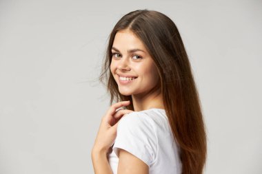 Uzun saçlı bir kadın açık arkaplanda gülümser ve elleriyle boynuna dokunur.