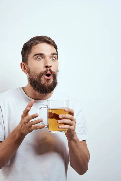 Hafif arka planda bira fincanı olan sakallı bir adam. Beyaz tişörtlü, alkollü bir içkinin görüntüsü. — Stok fotoğraf