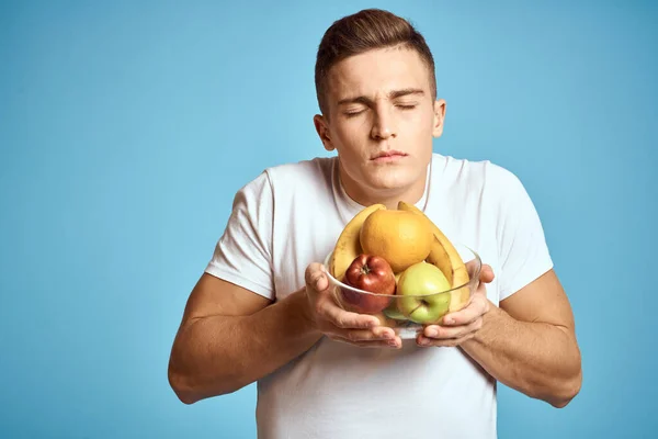 Hombre feliz con frutas frescas gesto con las manos fondo azul camiseta blanca vitaminas plátanos naranjas manzanas — Foto de Stock