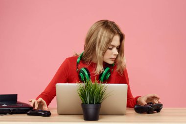 Kulaklık takan, dizüstü bilgisayarlı bir kadın masa başı eğlence sektöründe pembe arka planda oturuyor.