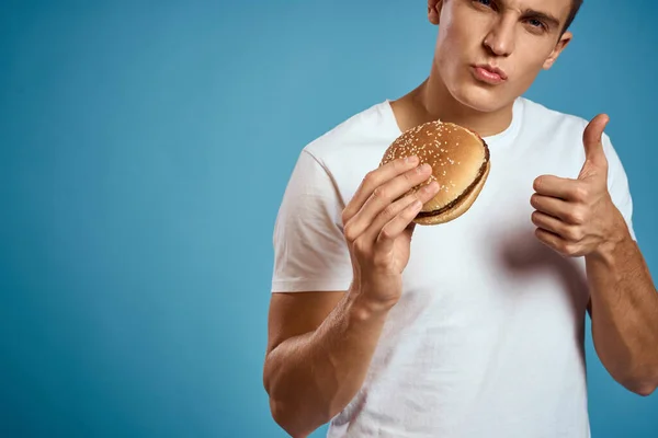 Mann mit Hamburger und weißem T-Shirt mit blauem Hintergrund gestikuliert mit Händen. — Stockfoto