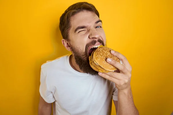 햄버거를 먹고 있는 남자가 패스트푸드인 화이트 티셔츠를 클로즈업 한 노란 배경의 남자 — 스톡 사진