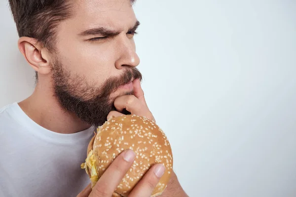 Um homem comendo um hambúrguer em um fundo leve em uma camiseta branca vista recortada close-up fome fast food — Fotografia de Stock