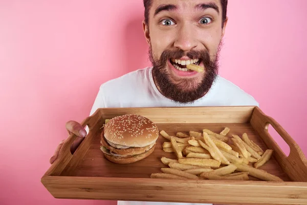Homem com uma bandeja de comida na mão hambúrguer batatas fritas e fast food caloria fundo rosa retrato close-up — Fotografia de Stock