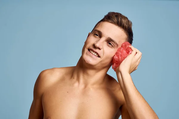 Молодой парень с розовой мочалкой в руке на синем фоне кожи уход дерматологии модели обрезанный вид — стоковое фото