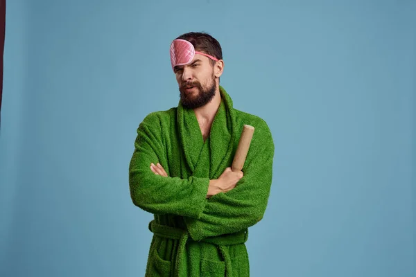Mężczyzna w różowej masce snu trzyma wałek w dłoni i zielony szlafrok niebieski tło emocje model — Zdjęcie stockowe